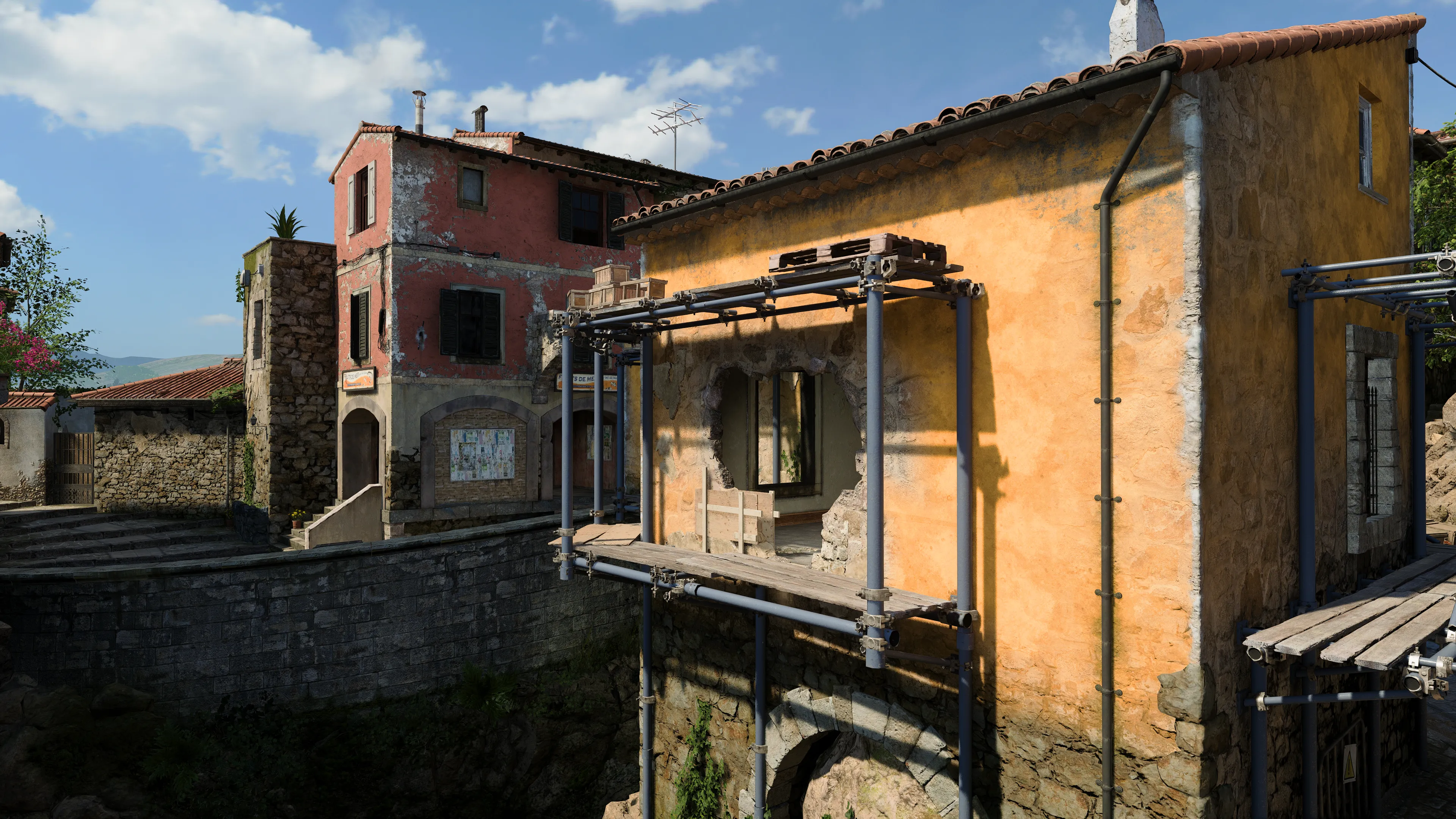 A weathered stucco building with scaffolding and a broken wall opening overlooks a small village square with colorful stone houses nearby.