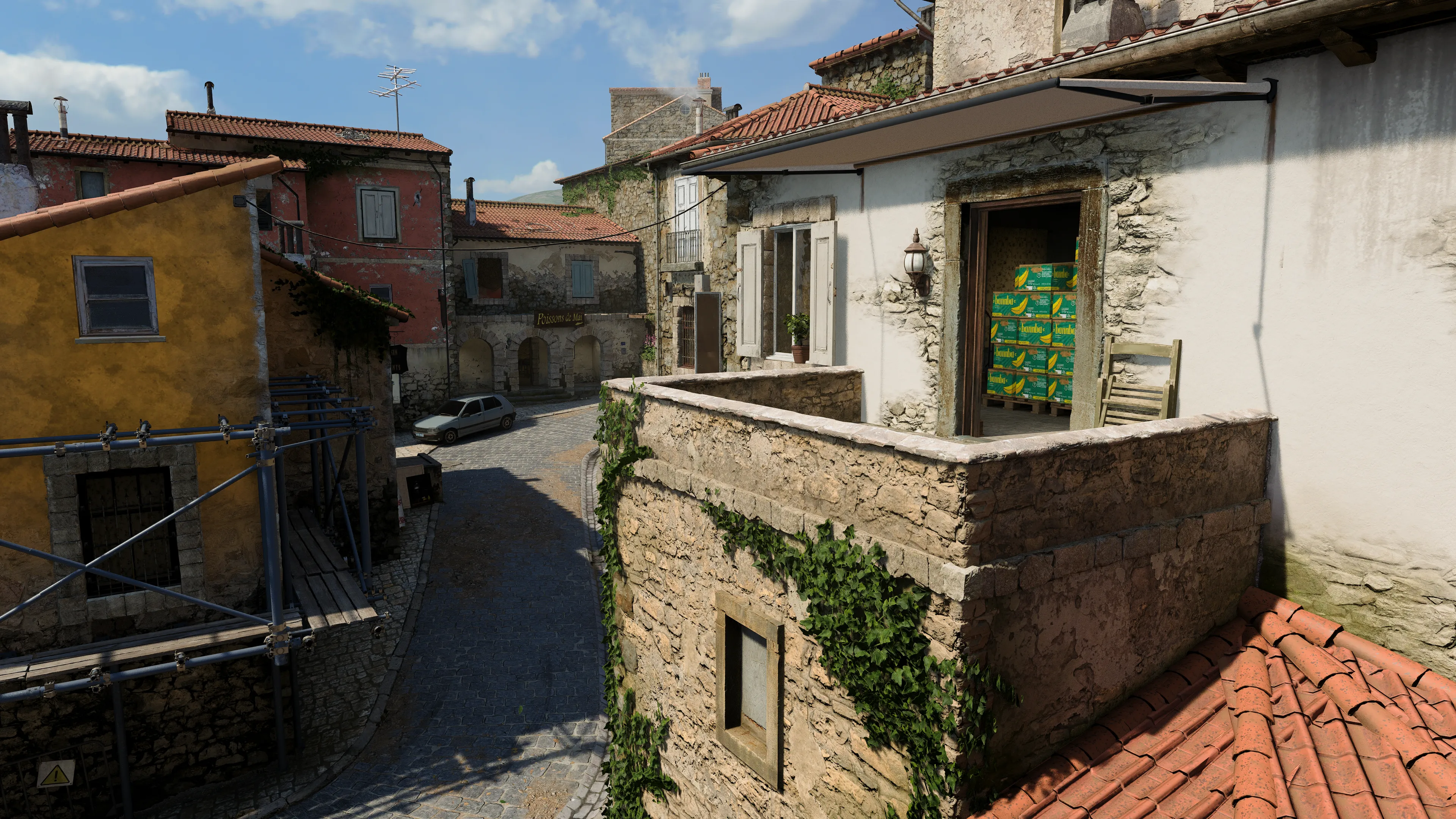 A narrow cobblestone street curves through a rustic village lined with stone houses and balconies, with an open doorway revealing stacked produce boxes inside.
