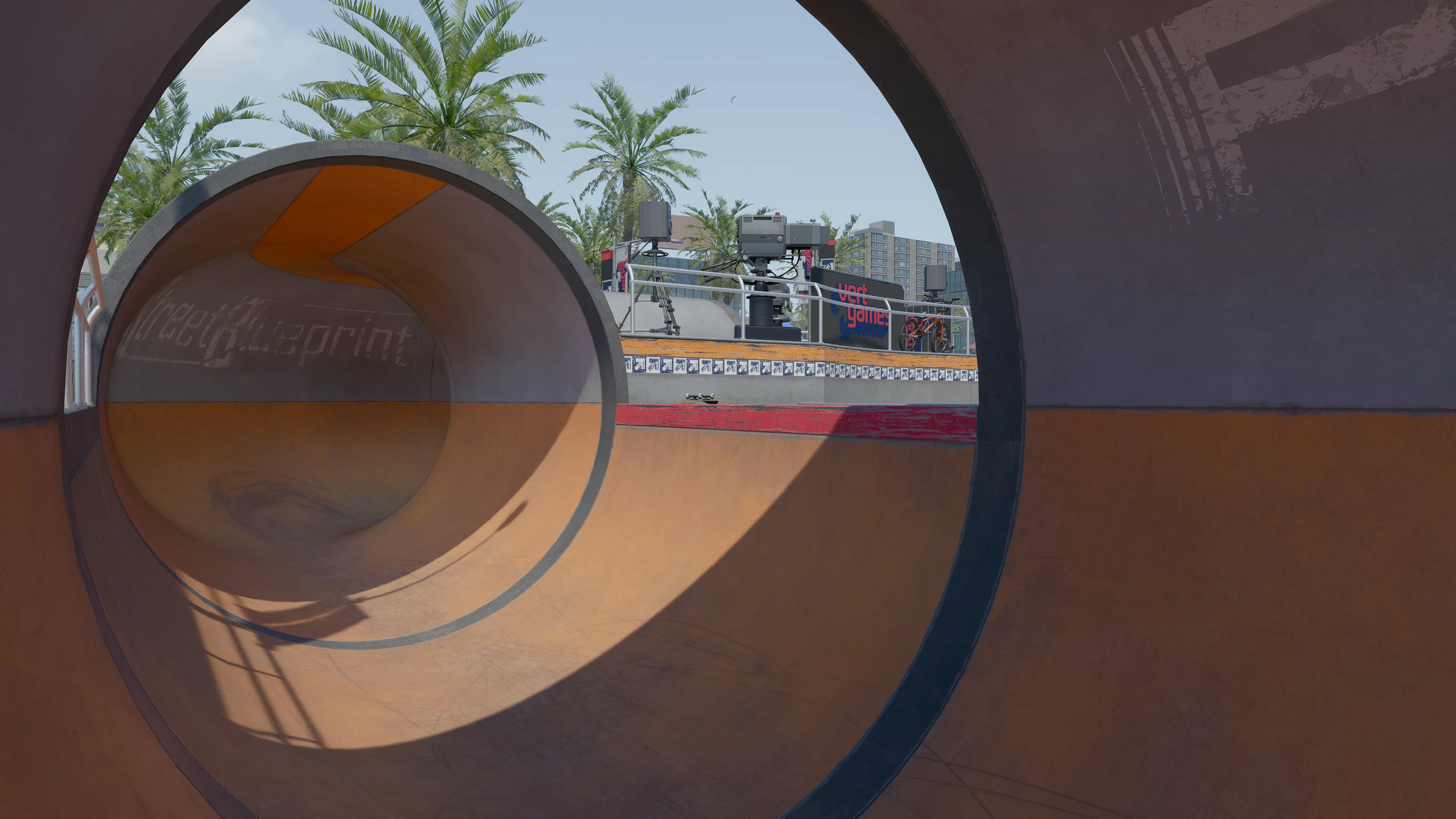 View through a circular skate pipe tunnel looking out toward the Vert Games skate park with palm trees and event equipment beyond.