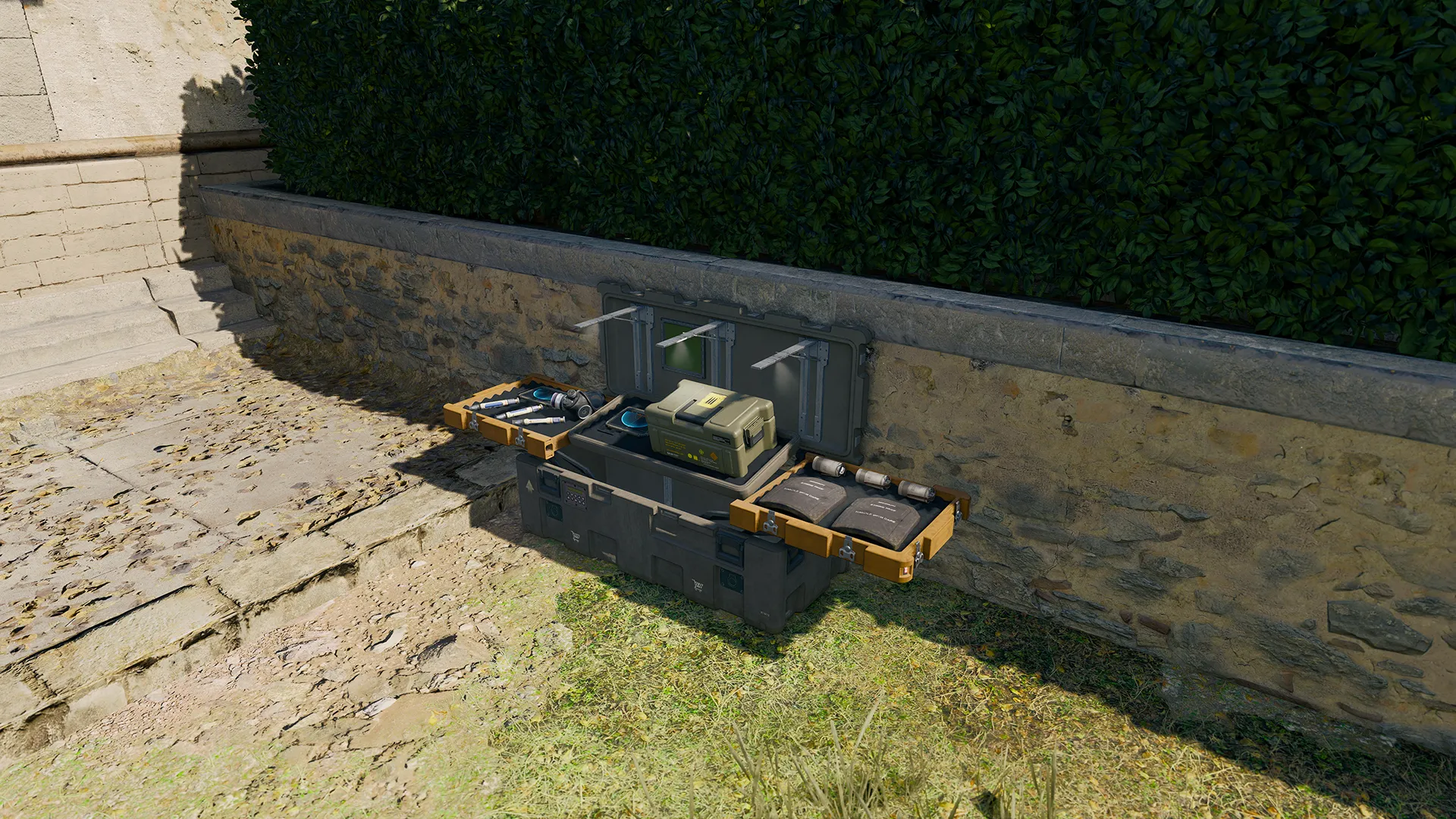Open equipment case with tactical gear placed beside a stone wall and hedge.